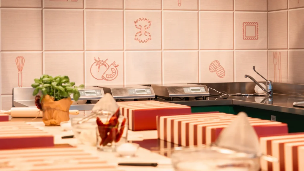 margherita_cooking_class-shoot_01-orizzontale-17 Postazioni individuali allestite per una cooking class italiana, con confezioni regalo a righe rosse e bianche, ingredienti freschi e utensili pronti all’uso in una cucina professionale sul Lago Maggiore.